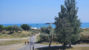 Ein Küstenpfad mit Spaziergängern, Bäumen und freiem Blick auf das Meer und Boote.