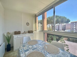 Ein Essbereich mit einem Tisch für zwei Personen, einem Fenster mit Blick auf Gebäude und Bäume sowie einem weißen Schrank mit Körben obenauf.