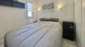 A bedroom with a bed, a window with blinds, a wall-mounted shelf, a wall-mounted light, and a small bedside table. - CAP D'AGDE - Plage Richelieu / Réf 193 (CAP D'AGDE)