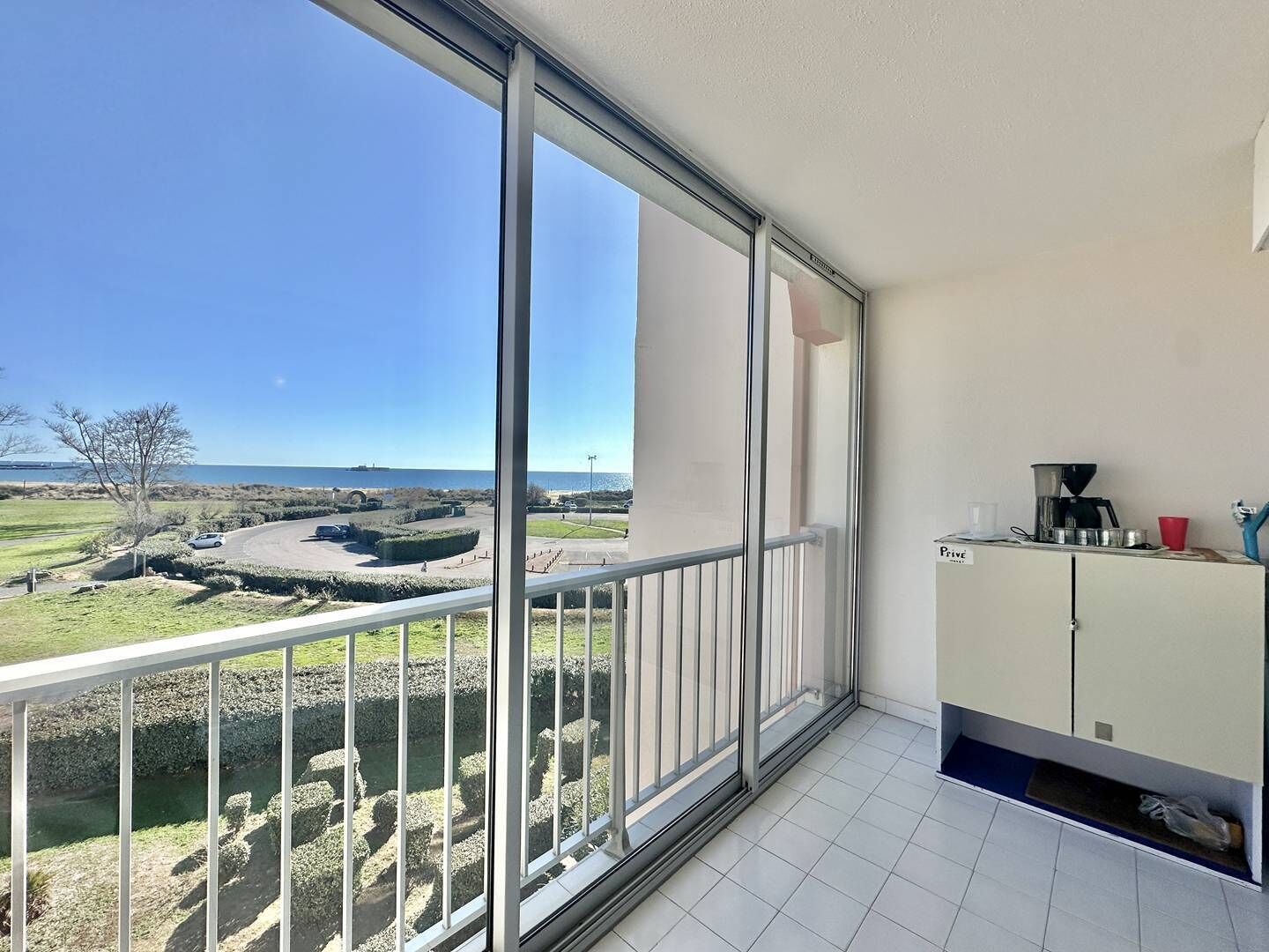 A balcony with a view of a coastal landscape, a kitchen area with a coffee machine, and a sign indicating a private room.