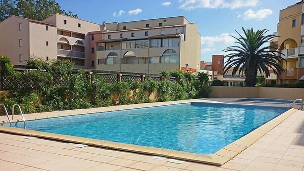 Una piscina rectangular con un borde de baldosas, rodeada de vegetación y edificios.