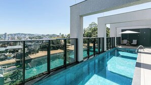 Una piscina en la azotea con barandillas de cristal, vistas de la ciudad y una zona de descanso con sillas y una sombrilla.
