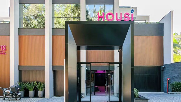 Un edificio moderno con un llamativo letrero de 'Housi', ventanas amplias y una entrada de vidrio.