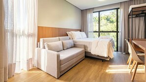 Habitación de hotel moderna con sofá, cama, escritorio y un amplio ventanal con vistas a zonas verdes.
