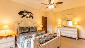 Quarto com cama, mesas de cabeceira, cômoda, ventilador de teto, espelho e uma decoração de urso na parede.