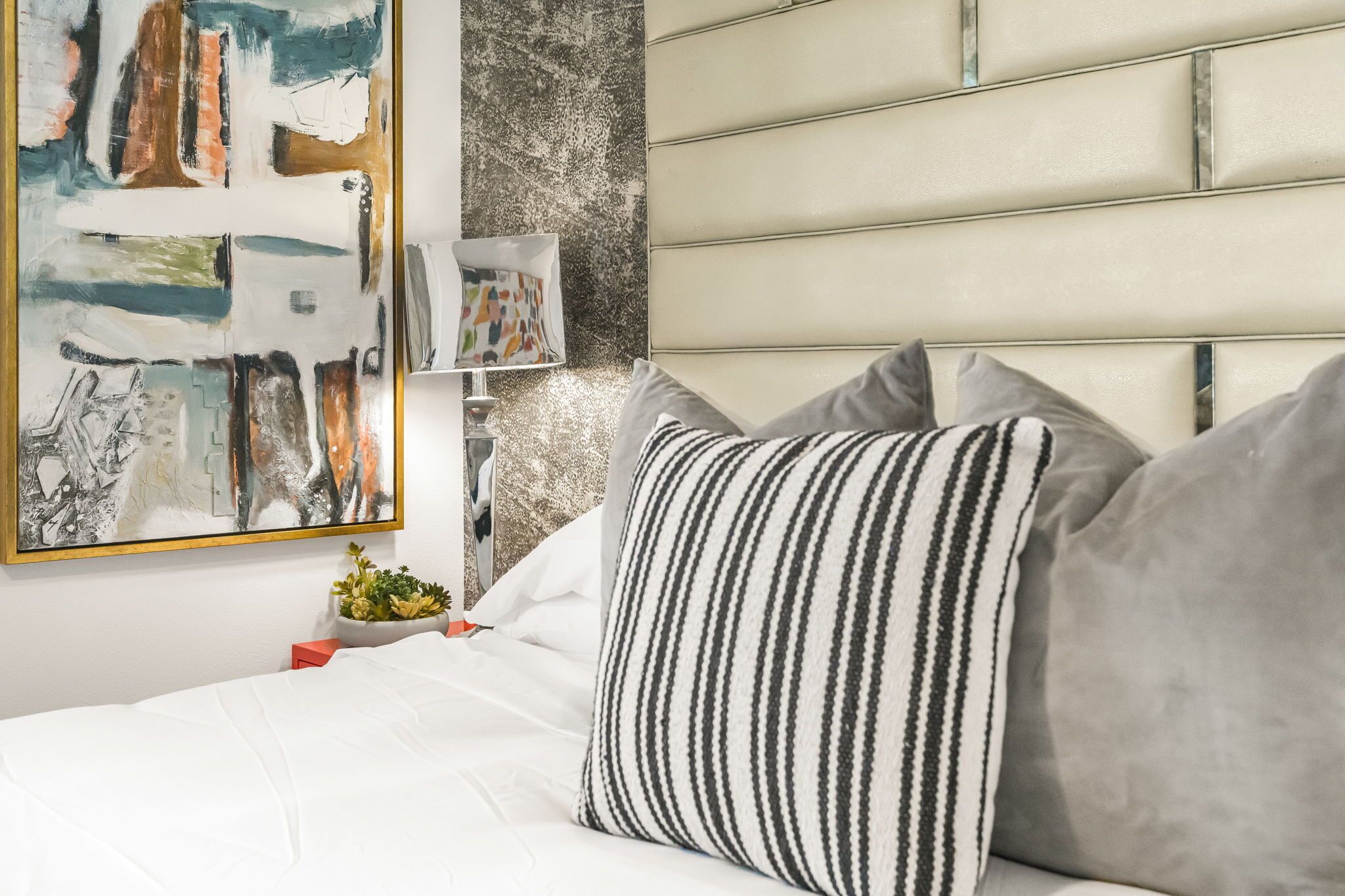 A modern bedroom with a headboard, a bed with white bedding, striped pillows, a small plant, and a colorful abstract painting.