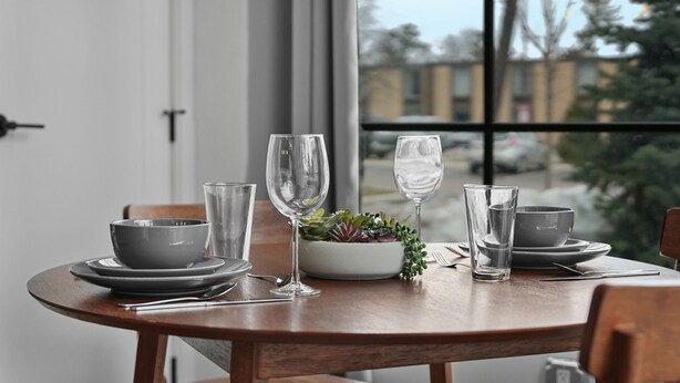 Een gedekte eettafel met borden, kommen en glazen, een saladeschaal en een raam met uitzicht op gebouwen en bomen.