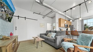 Een moderne woonkamer met een bank, salontafel en stoelen, een keuken met kastjes en apparatuur, en een groot raam.