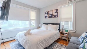 Une chambre à coucher avec un lit, des tables de chevet, une télévision et une fenêtre avec des stores.