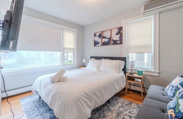 A bedroom with a bed, bedside tables, a TV, and a window with blinds.