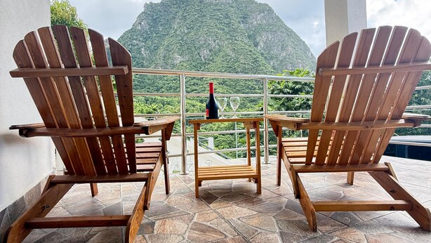 Two wooden Adirondack chairs facing a table with a bottle and glass, on a balcony with stone flooring and a view of a mountain. - Tranquil & private 1-bedroom fully furnished AC apartment in charming Soufrière (Soufrière)