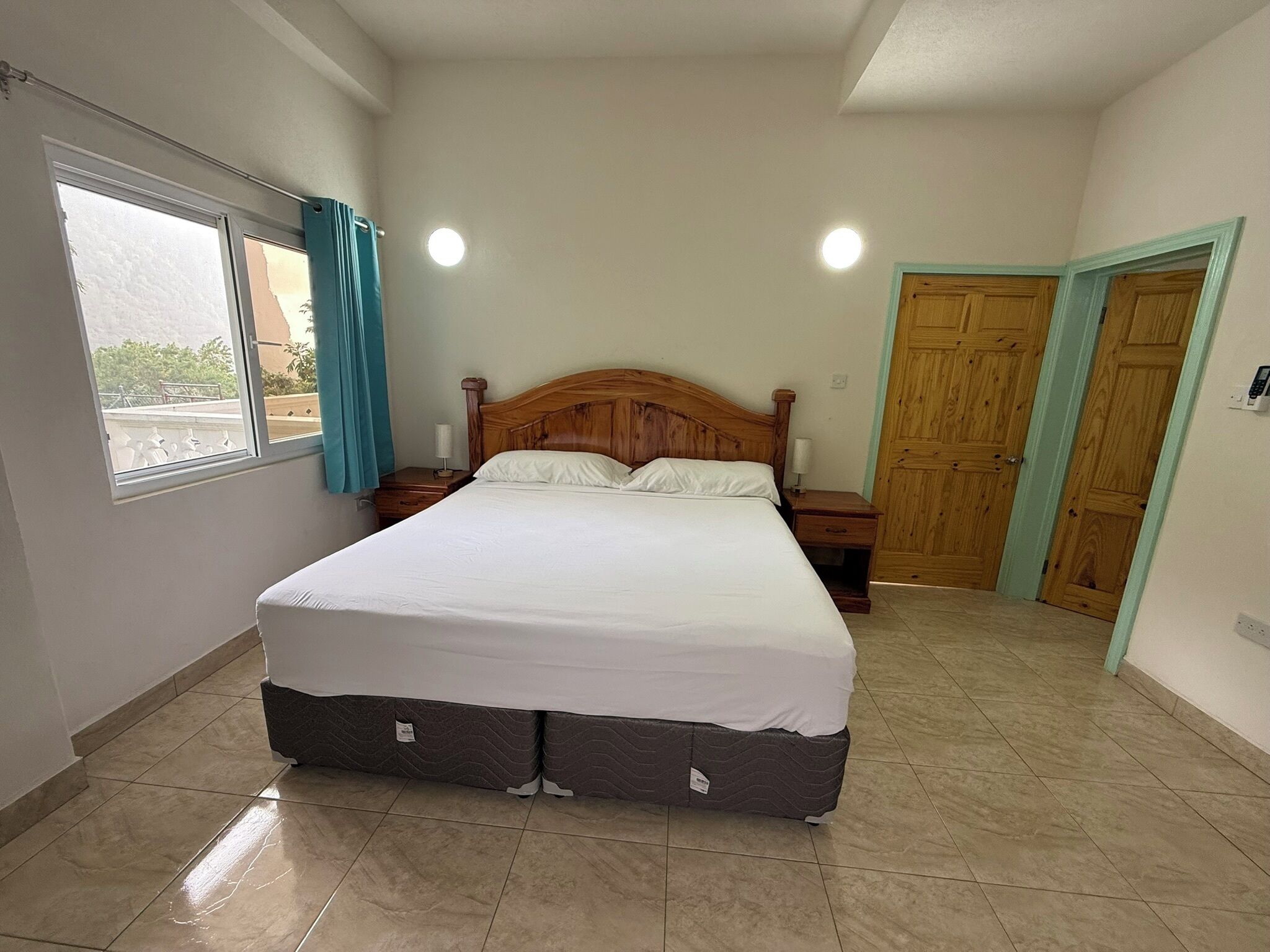 Une chambre à coucher avec un grand lit, une fenêtre avec des rideaux, une table de chevet, une porte en bois et un plancher carrelé.