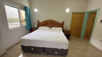 Une chambre à coucher avec un grand lit, une fenêtre avec des rideaux, une table de chevet, une porte en bois et un plancher carrelé.