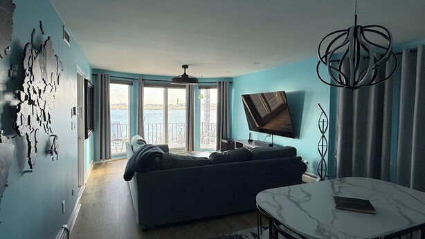 A living room with a sofa, a TV, a dining table, and a view of the sea through large windows.
