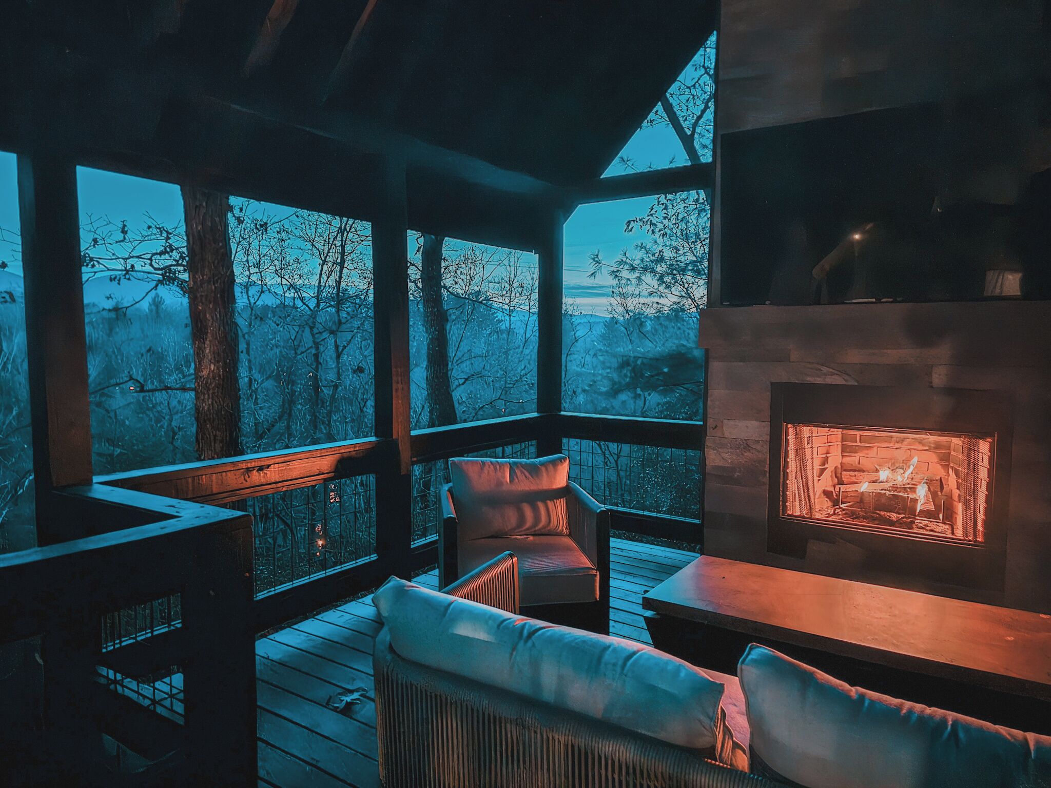 A cozy outdoor seating area with a fireplace, wooden deck, and a view of trees.