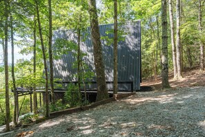 A modern building with a metal exterior, surrounded by a forested area with a gravel path. - Luxe A-frame Hideaway, mountain views, hot tub, outdoor patio w/fireplace (Blairsville)