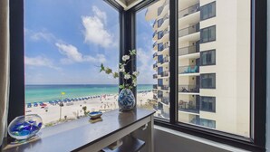 Une vue sur une plage, prise depuis un balcon, avec un vase de fleurs et un saladier en verre posés sur le rebord de la fenêtre.