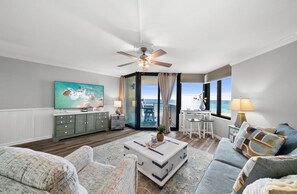 A spacious living room with a large television, a ceiling fan, and a view of the ocean through the glass door. - Sunbird Shores 511E-Gulf Front-Sleeps 4 (Panama City)