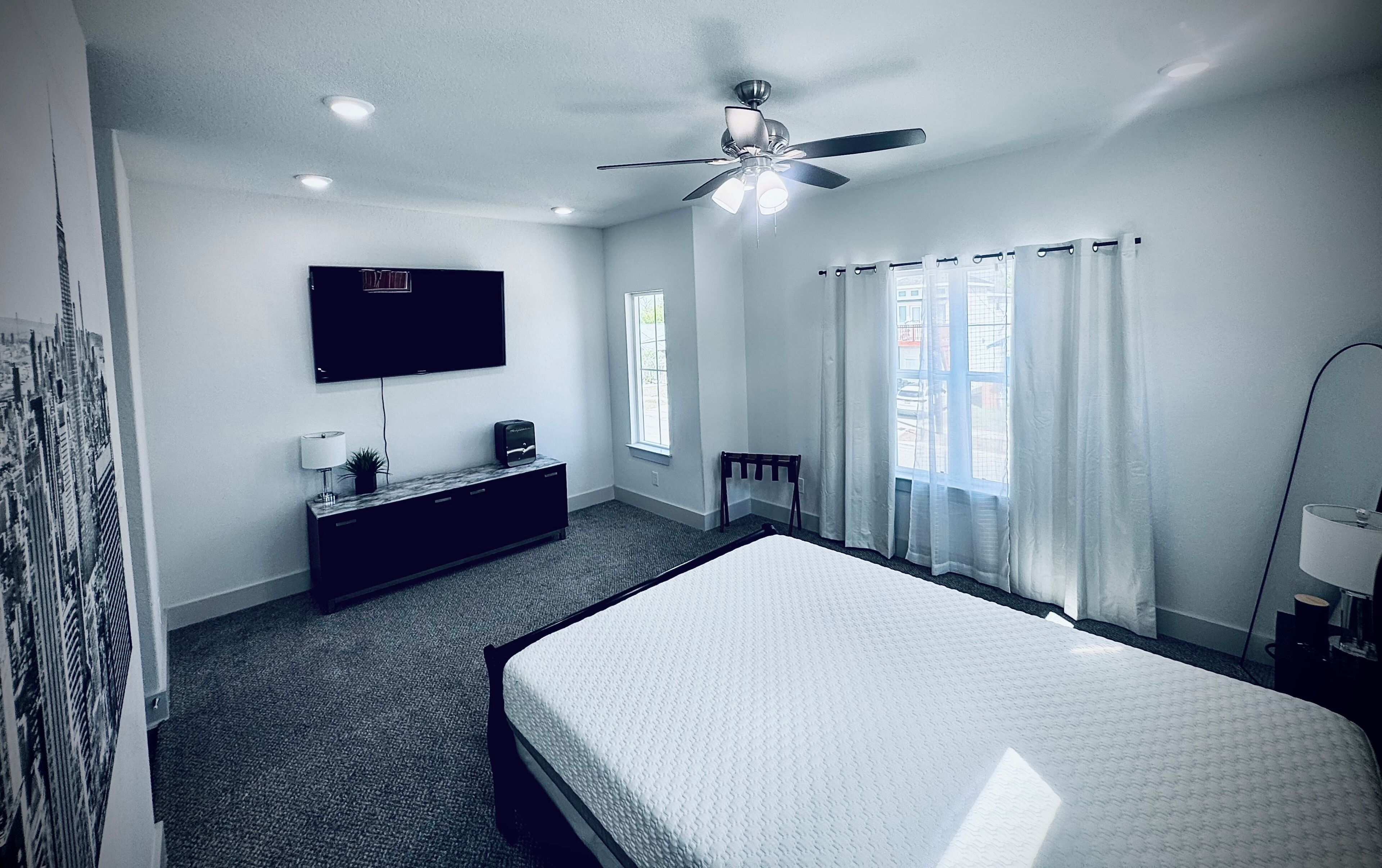 A hotel room with a bed, a TV, a ceiling fan, a window with curtains, and a small table with a lamp.