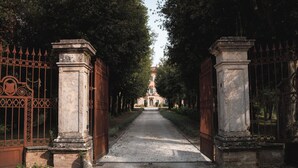 A pathway leading to a building with a central tower, flanked by ornate iron gates and stone pillars. - Cosy apartment on a tranquil estate with shared pool & mountain views (Marche)