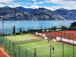 A tennis court with a net, surrounded by a fence, with mountains and a lake in the background. - Apartment Blue by Interhome (Albisano)