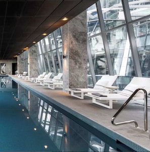A modern indoor swimming pool with clear glass walls, white lounge chairs, and a tiled floor. - Royal Executive Living  Sky Corporate Residence (Melbourne)