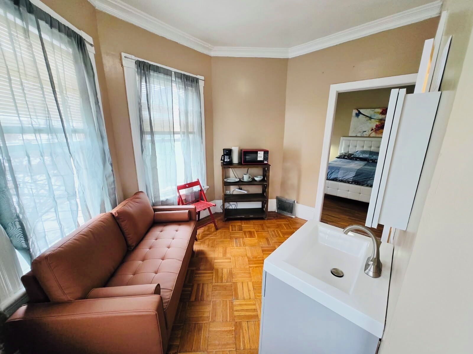 A compact room with a brown sofa, a white sink, a bed with a blue bedspread, and a small shelf with a microwave and cups.