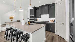 A modern kitchen with a large island, dark cabinets, stainless steel appliances, and pendant lighting. - Sun-Filled Coastal Home | Lagoon Access| Hot Tub| Sleeps 12 (St. George)