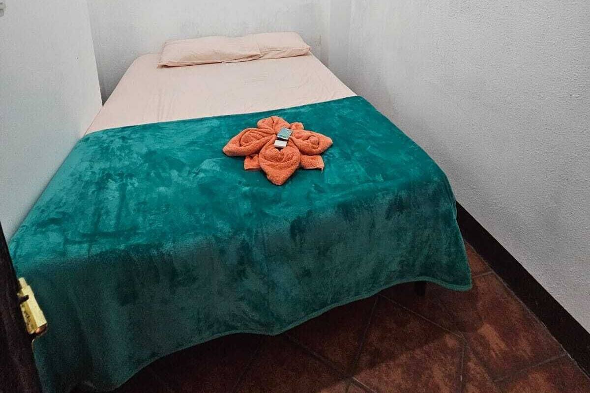 Ein kleines Zimmer mit einem Einzelbett, einer türkisblauen Bettdecke und einem Handtuch, das wie eine Blume auf dem Bett drapiert ist.