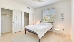 Une chambre à coucher comprenant un lit, un ventilateur de plafond, une fenêtre avec des stores, un placard et une petite table ronde.