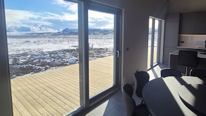 Un interior moderno con un gran ventanal corredera que da a una terraza de madera, un comedor con sillas y una cocina con armarios.