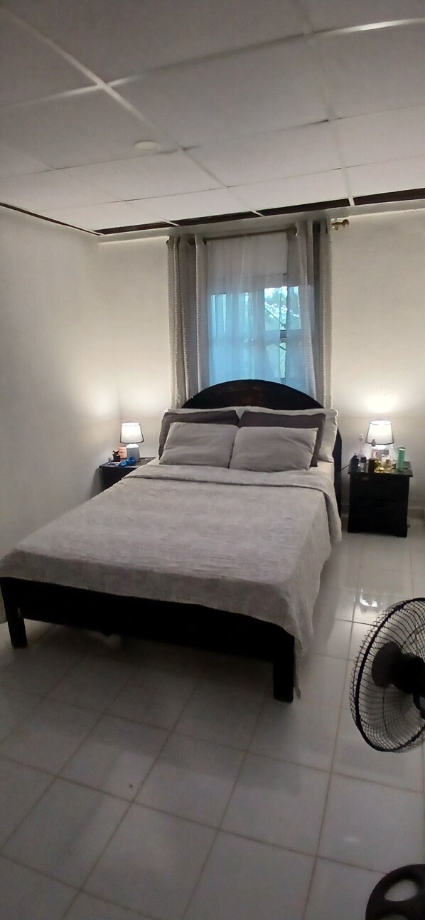 A bedroom with a bed, two bedside tables with lamps, a ceiling fan, and a window with curtains.