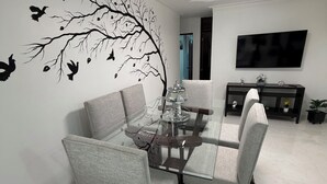 Zona pranzo con tavolo in vetro, sedie rivestite di grigio, un murale raffigurante un albero con degli uccelli, una ventola a soffitto e un televisore montato a parete.