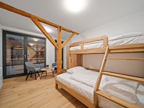 Een slaapkamer met stapelbedden, een houten balkenplafond, een klein tafeltje met stoelen en uitzicht door een raam met jaloezieën.