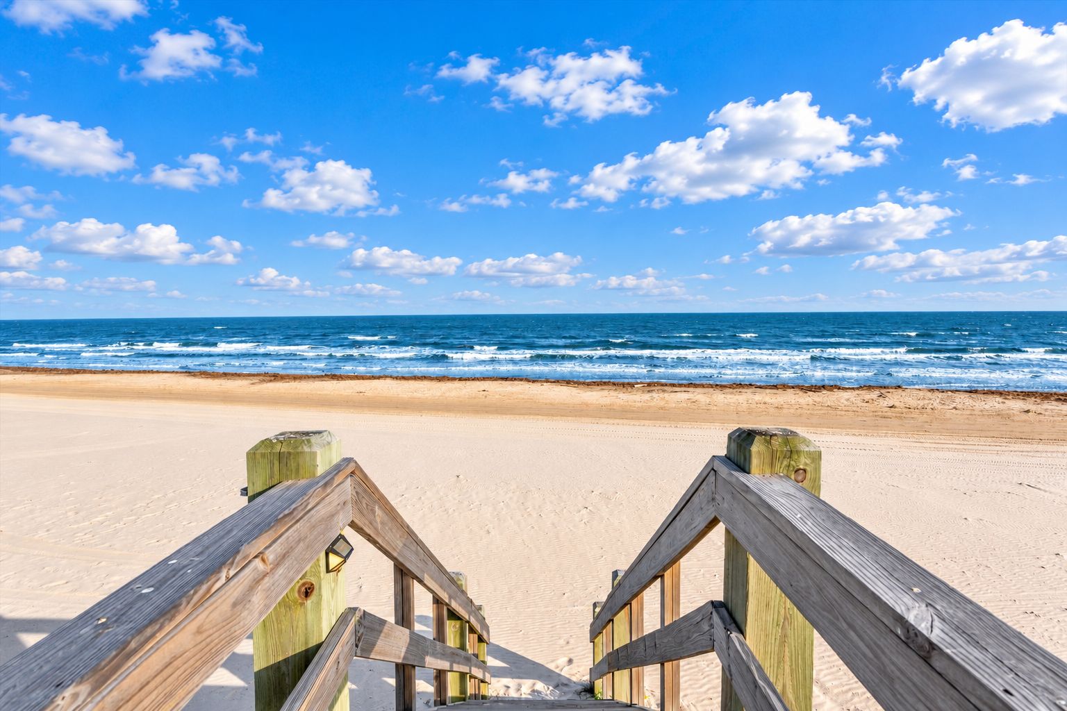 Een houten looppad dat naar een zandstrand leidt, met de oceaan op de achtergrond.