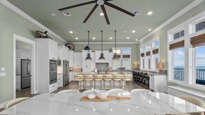 A spacious kitchen with white cabinets, a central island with stools, and a dining area with a large table and chairs. - Beachfront Retreat Sleeps 30 with Swim Spa, Fire Pit and Putt-Putt (Bolivar Peninsula)