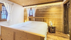Ein Schlafzimmer mit Holzboden, einem Bett, einem Fenster mit Vorhängen, einem Nachttisch mit Lampe und einer Holzwand.