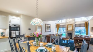 Een moderne keuken met dubbele ovens, een eetgedeelte met een houten tafel gedekt voor vier personen, en een woonkamer met een open haard en uitzicht op de omgeving.
