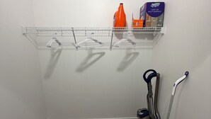 Esquina de una habitación con un estante blanco, un aspirador y una escoba.