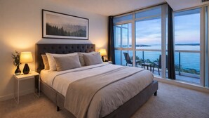 A bedroom with a large bed, two bedside tables with lamps, a framed picture on the wall, and a view of the sea through large windows. - 2 Bedroom Luxury Apartment in Downtown Toronto (Toronto)