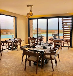 Un comedor con mesas y sillas, ventanas amplias con vistas a una piscina y paisajes, y lámparas colgantes.