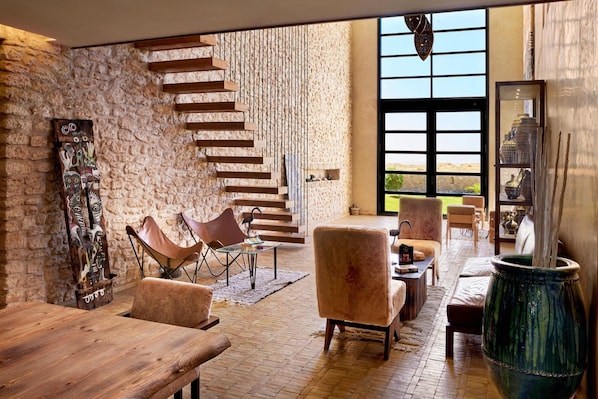 Un amplio salón con pared de piedra, escalera de madera, sillones de cuero y un ventanal con vistas.