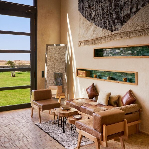 A spacious living area with a large window, a stone wall, a fireplace, and a wooden coffee table. - Baberrih (Moulay Bouzarqtoune)