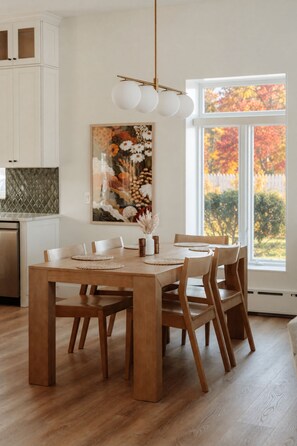 Un comedor con una mesa de madera, sillas, una lámpara colgante y un ventanal con vista a árboles.