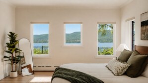 A bedroom with a large bed, a potted plant, a framed picture, and three windows offering a view of a lake and hills.