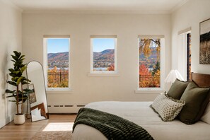 Un dormitorio con una cama grande, tres ventanas, una planta en maceta y un cuadro en la pared.