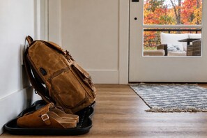 Una mochila marrón apoyada contra una pared, un par de zapatos marrones sobre un tapete negro, y vista de un porche con una silla de mimbre y un cojín blanco.
