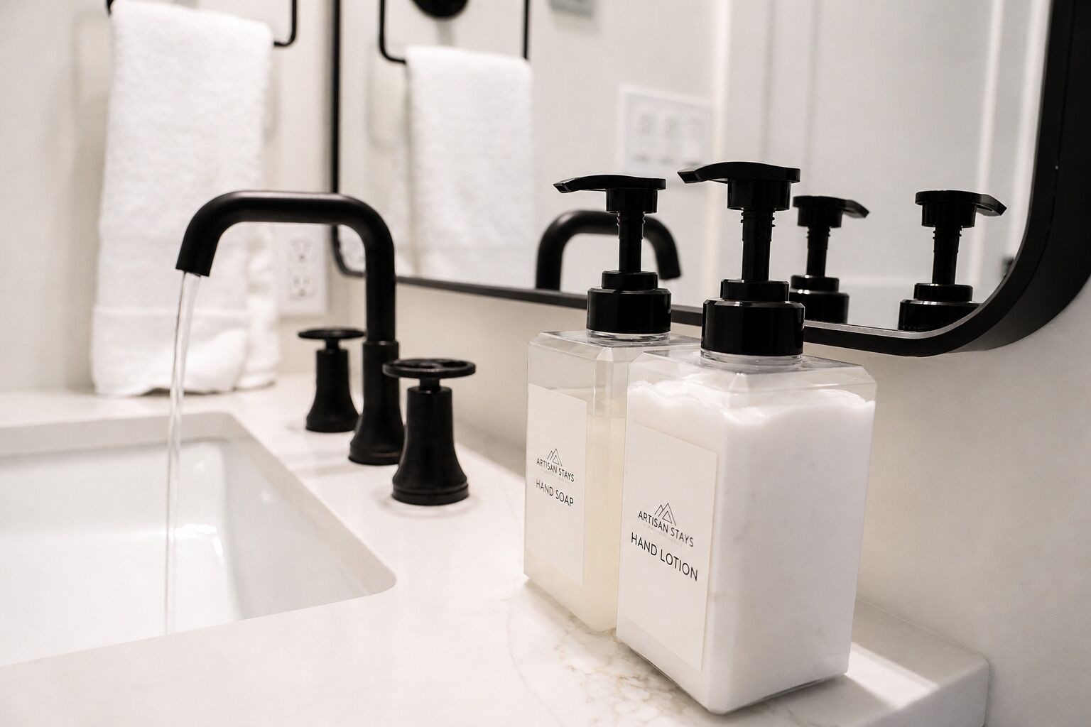 Un baño con lavamanos blanco, grifos negros y dos botellas de jabón de manos y loción con la etiqueta 'Artisan Stays'.