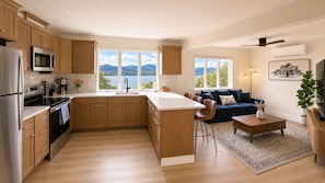 A modern kitchen with wooden cabinets, stainless steel appliances, and a large island. There is a dining area with a blue sofa and a coffee table. The room has large windows with a view of mountains and a lake.