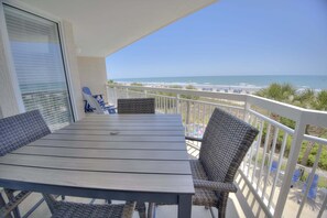 Varanda com mesa e cadeiras, vista para uma praia com barcos e guarda-sóis.